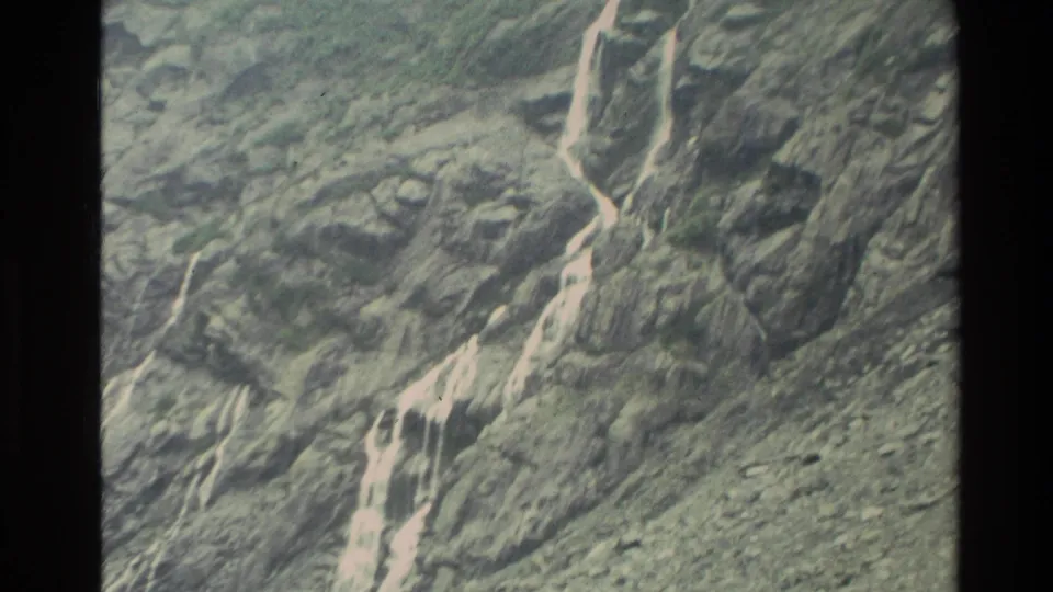 1982 New Zealand 8mm frame of a tall segmented waterfall flowing over a rugged gray-green mountain slope.