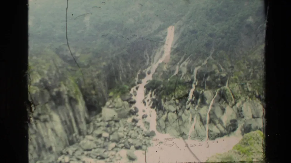 1982 New Zealand 8mm frame of a pale creek channel cutting through rocks and green-gray mountain terrain.
