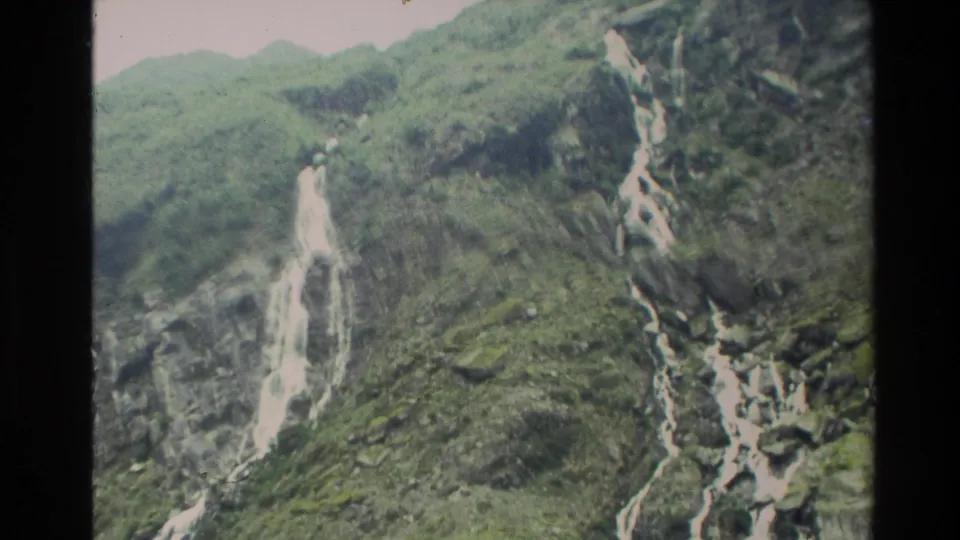1982 New Zealand home-movie frame with tall waterfalls cutting through a green mountain slope.
