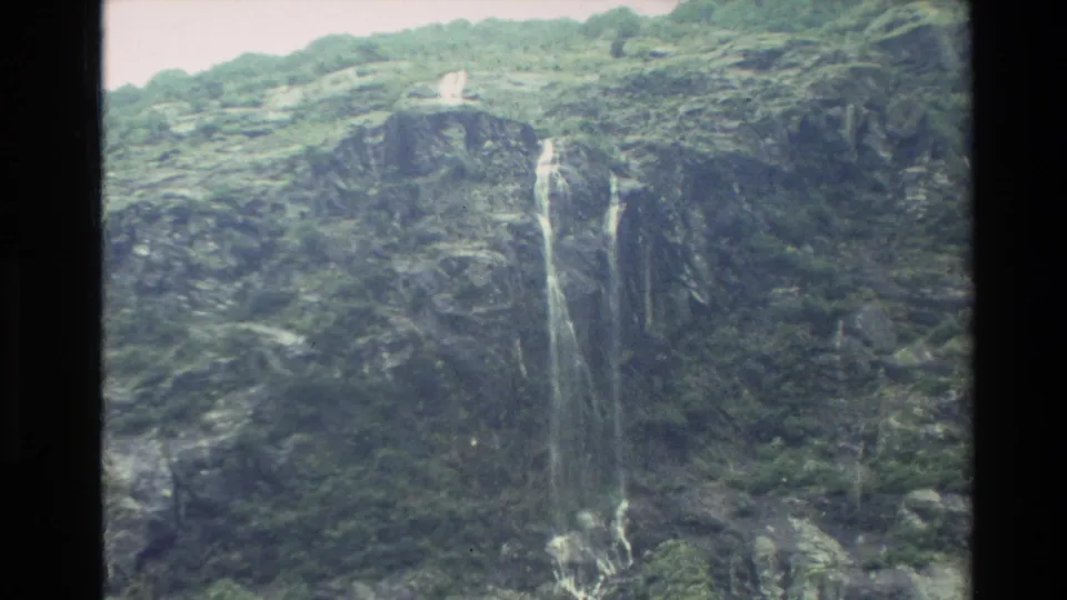 1982 New Zealand 8mm frame showing narrow waterfalls falling down a shadowed cliff wall.