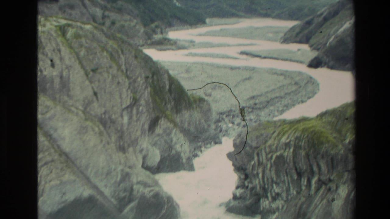 1982 New Zealand 8mm frame showing a pale braided river below steep mossy canyon slopes and gray rock formations.