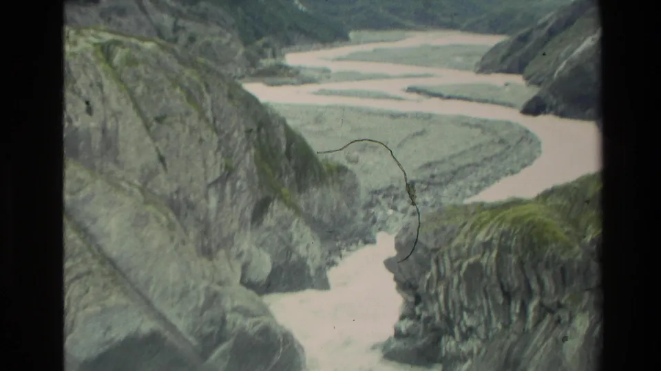 1982 New Zealand 8mm frame showing a pale braided river below steep mossy canyon slopes and gray rock formations.