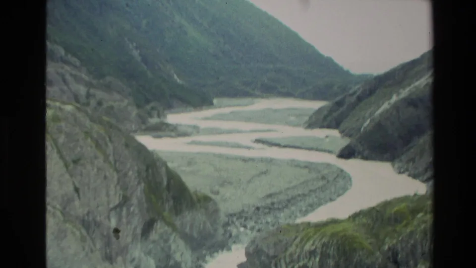 1982 New Zealand home-movie frame with a wide view of pale river channels between steep green and gray canyon sides.