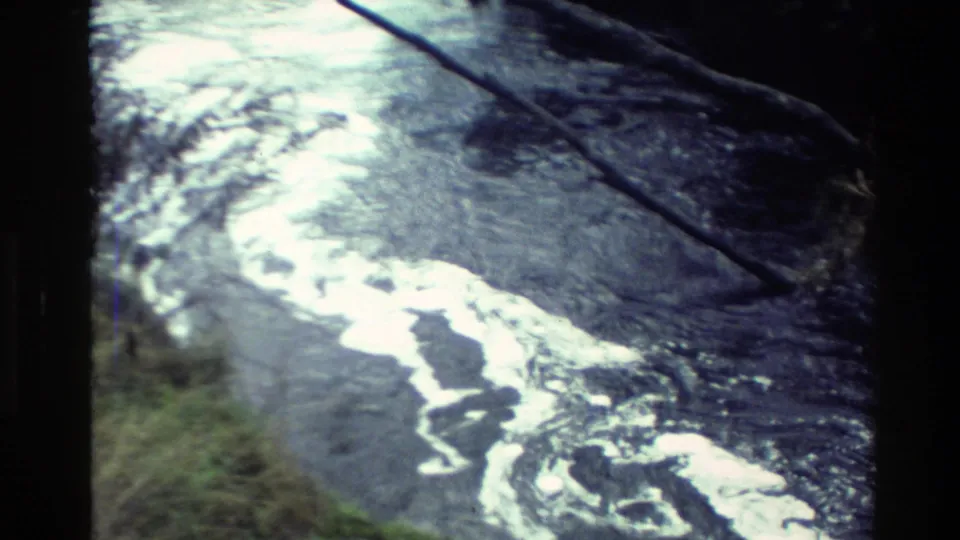 1982 New Zealand 8mm frame showing rippled stream water, foam trails, and an overhanging branch.