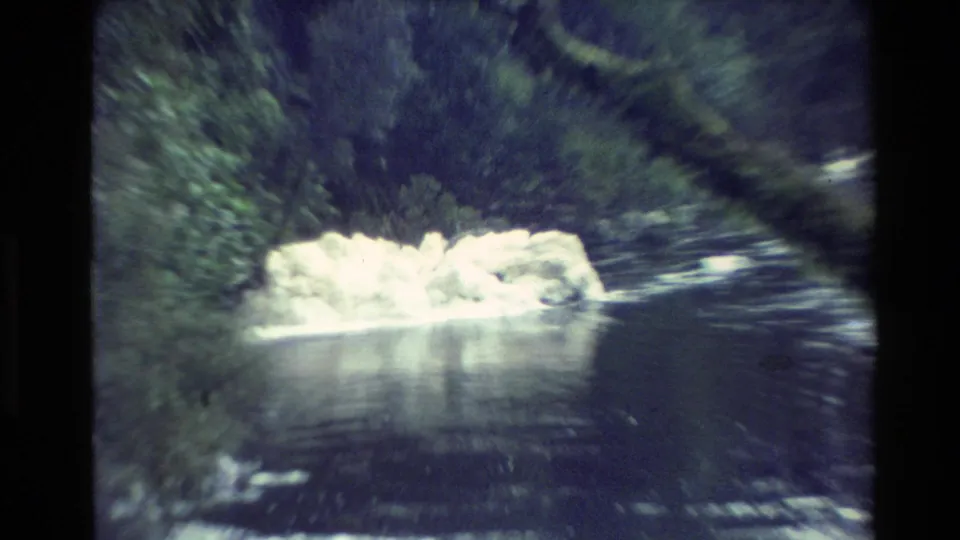 1982 New Zealand 8mm frame of a bright ice formation lodged in a flowing river near a wooded bank.