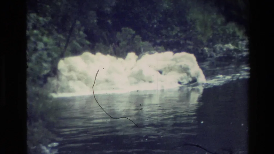 1982 New Zealand 8mm frame showing a broad white ice mass against reflective river water and dense riverside trees.