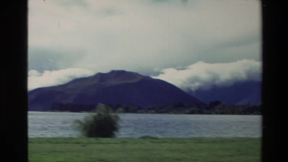1982 New Zealand 8mm frame looking across a lake toward dark mountains partly covered by low cloud.
