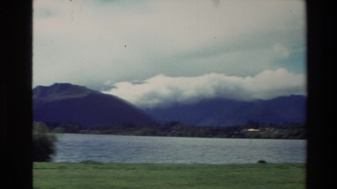 1982 New Zealand 8mm view of a lake, grassy shore, and cloud-covered mountains seen through a dark window frame.