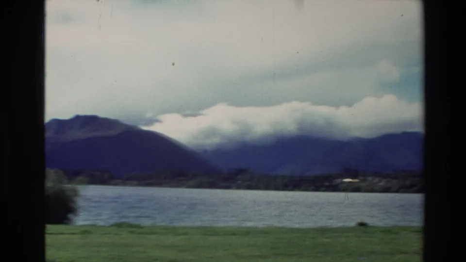 1982 New Zealand 8mm view of a lake, grassy shore, and cloud-covered mountains seen through a dark window frame.