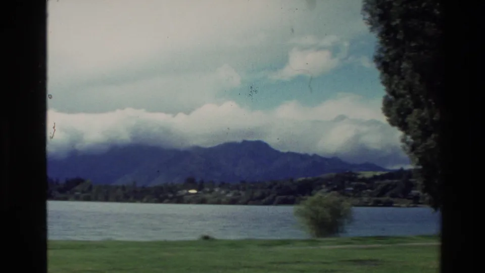 1982 New Zealand home-movie frame showing a lake, green shore, small tree, and mountains partly covered by clouds.