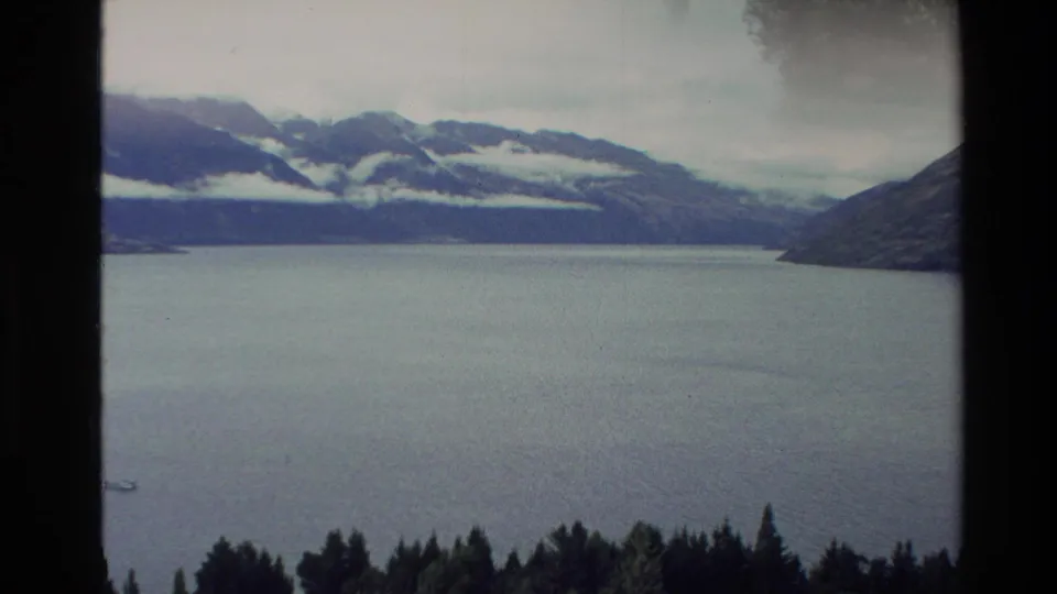 1982 New Zealand 8mm frame looking across a broad lake toward mountains and low cloud, with dark window framing at the sides.