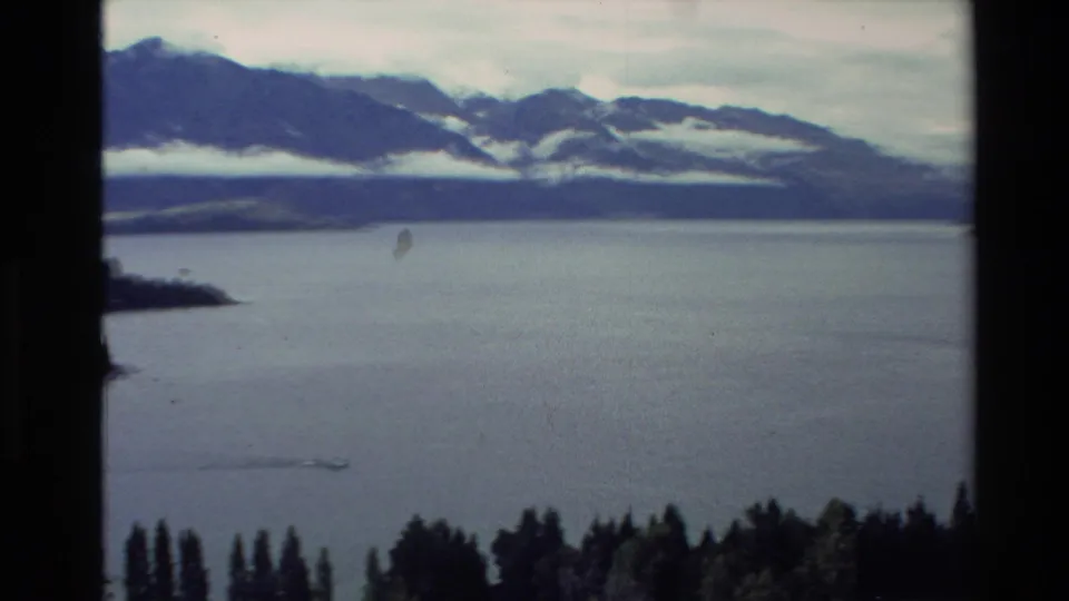 1982 New Zealand home-movie frame with a lake, tiny distant boat, foreground trees, and cloud-banded mountains.
