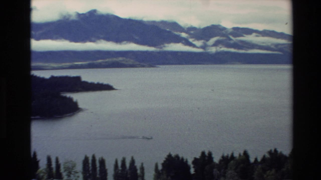 1982 New Zealand 8mm frame showing a calm lake below misty mountains, with forested shoreline and foreground trees.