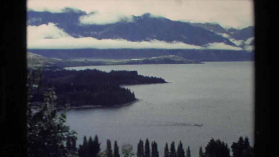 1982 New Zealand 8mm view of a wooded peninsula on a lake, with misty mountains and a small boat crossing the water.
