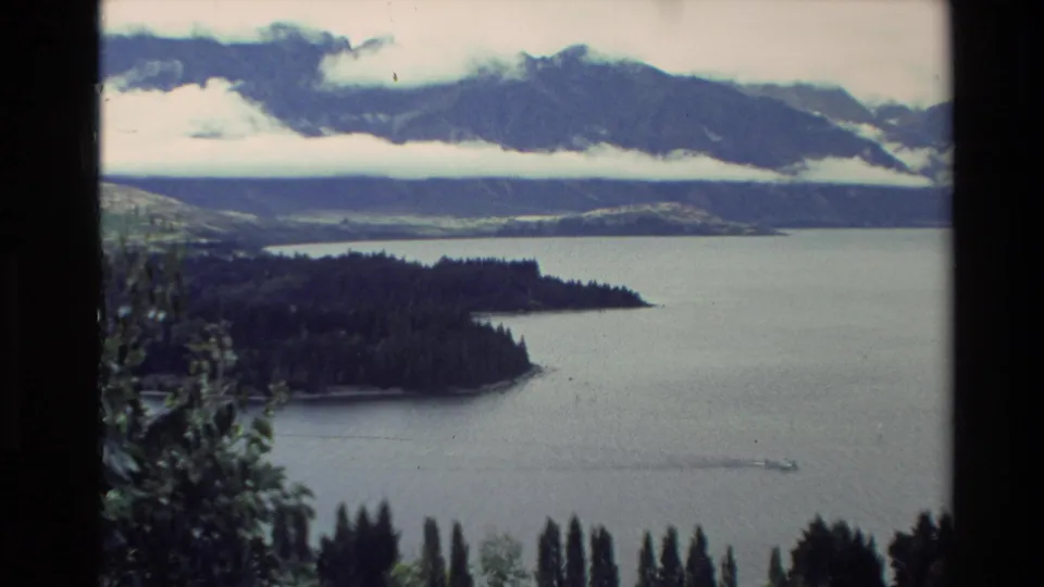 1982 New Zealand 8mm frame of a forested lake peninsula, distant misty mountains, and a small boat moving across the water.