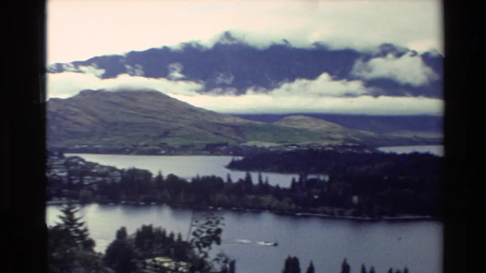 1982 New Zealand 8mm panorama of a lake below mountains, with shoreline trees, buildings, and a small boat wake.
