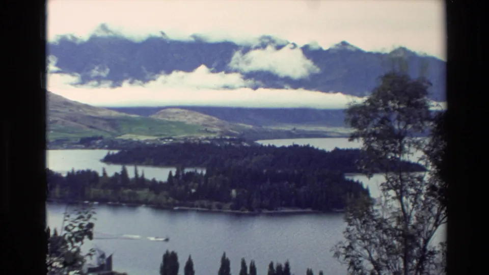 1982 New Zealand scenic overlook with lake water, a forested peninsula, distant mountains, low clouds, and foreground branches.