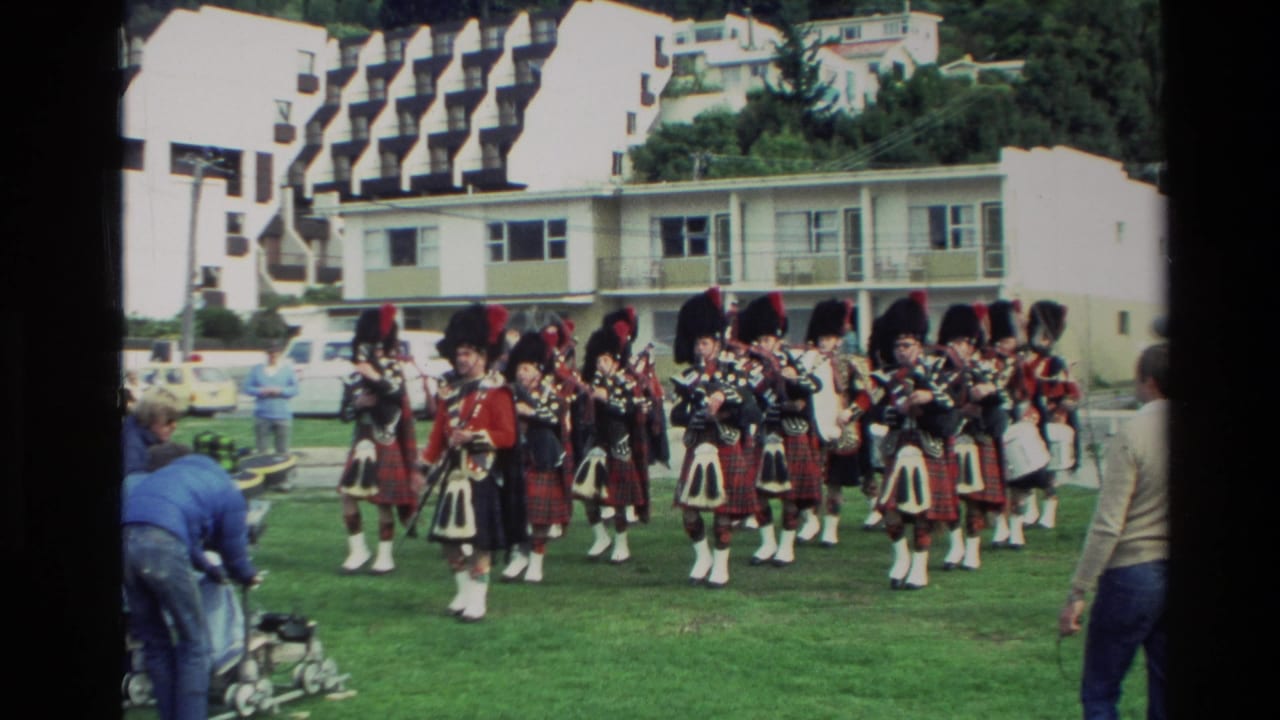 1982 New Zealand 8mm frame showing a Scottish pipe band in tartan uniforms performing on grass beside resort-like white buildings.