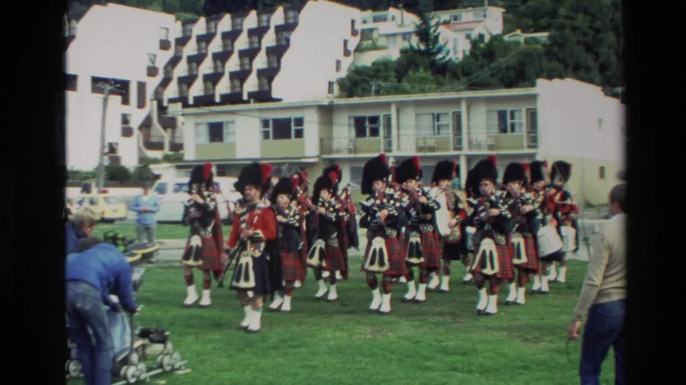 1982 New Zealand 8mm frame showing a Scottish pipe band in tartan uniforms performing on grass beside resort-like white buildings.