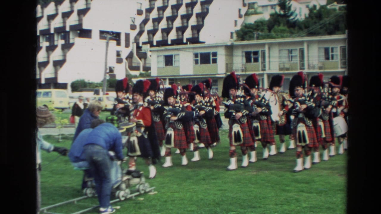 Pipe band in tartan uniforms stands in formation as a camera operator works beside the lawn.