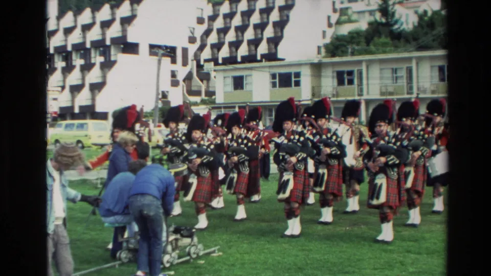 A Scottish pipe band marches and plays on a lawn in 1982 New Zealand, with camera equipment and spectators at the edge of frame.