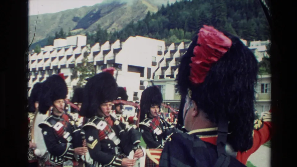 Close 8mm view of Scottish pipe band musicians in black feathered hats and tartan uniforms, with New Zealand hills in the background.
