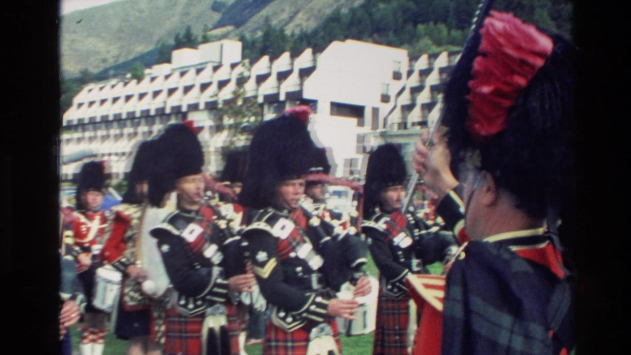 1982 New Zealand home-movie frame showing Scottish pipe band members in tartan kilts and dark feather bonnets preparing or playing during a parade.