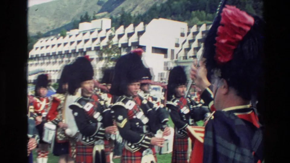 1982 New Zealand home-movie frame showing Scottish pipe band members in tartan kilts and dark feather bonnets preparing or playing during a parade.