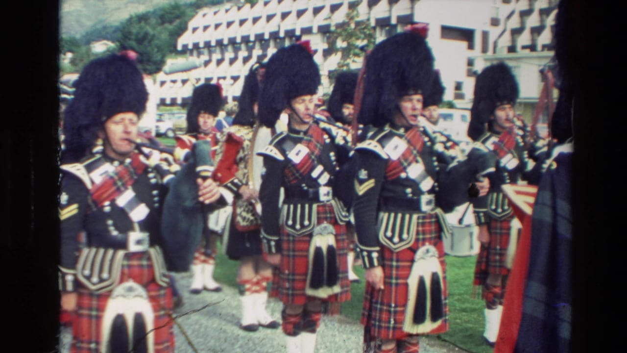 Bagpipe players and a drummer in red tartan uniforms stand in formation outdoors.