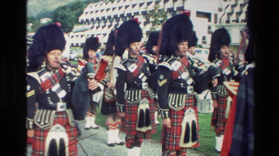 Scottish pipe band members in ceremonial dress play bagpipes and drums in an outdoor New Zealand parade setting, with spectators partly visible.
