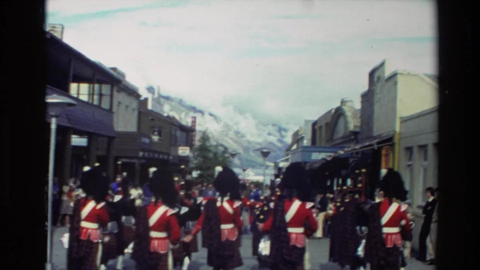A 1982 New Zealand parade view from behind shows the pipe band marching along a street lined with spectators, with mountains beyond the buildings.