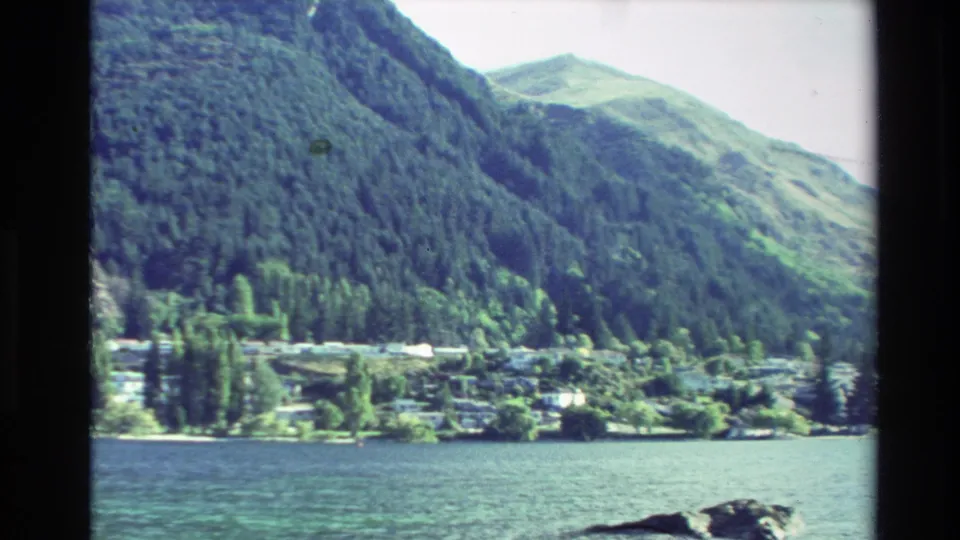 1982 New Zealand lakeside view with rippled water, distant houses, shoreline trees, and a large forested hillside.