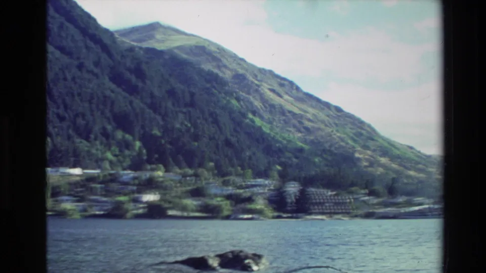 1982 New Zealand 8mm frame with motion blur across water, clustered shoreline buildings, trees, and sloping green hills.