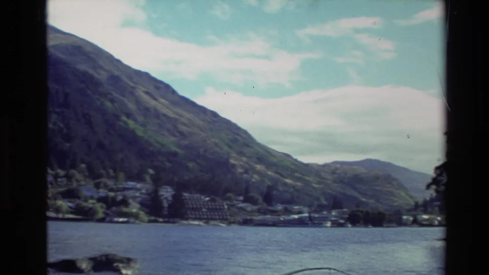 1982 New Zealand travel frame showing open water, a distant lakeside town, steep hills, and partly cloudy sky.
