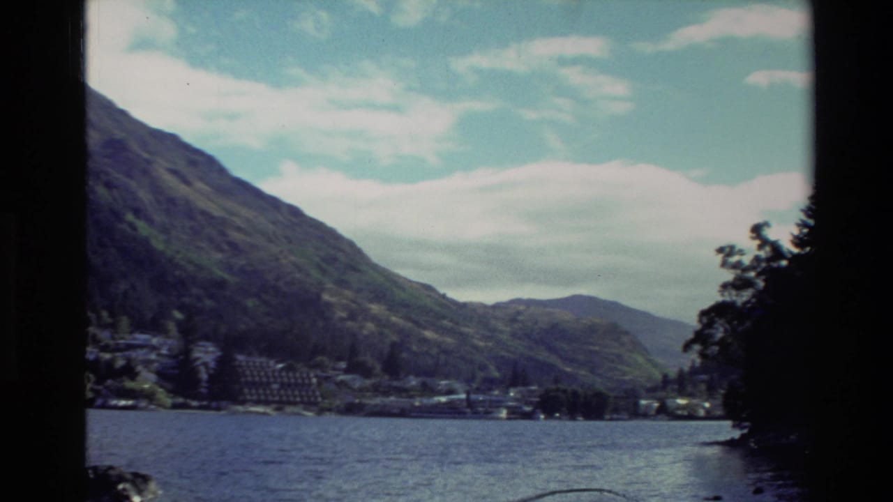 1982 New Zealand home-movie view of a calm lake, forested mountain slopes, and a small hillside town under pale clouds.