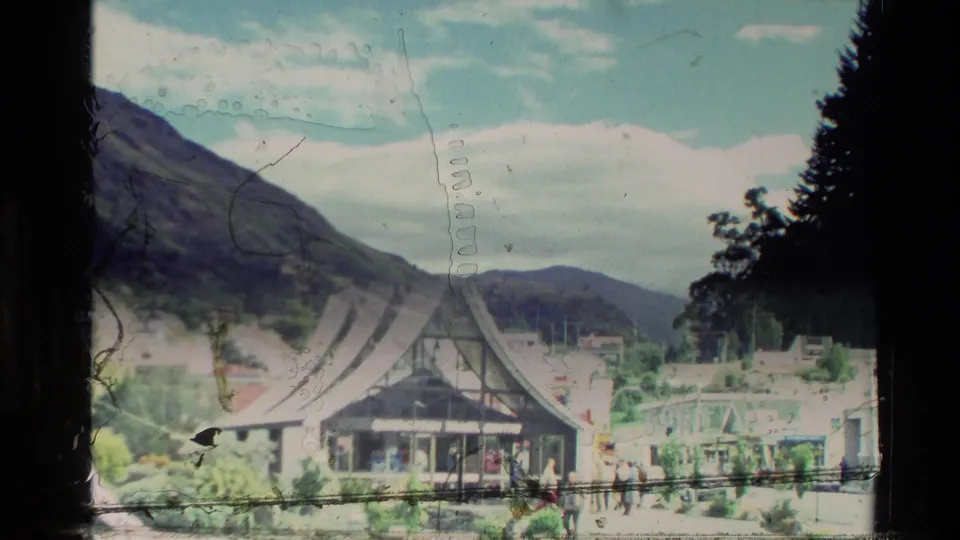 1982 New Zealand travel frame showing a distinctive steep-roofed building below forested mountains, with small distant people nearby.