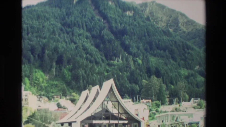 1982 New Zealand home-movie frame of a sharply roofed building below a green forested mountainside and nearby town structures.
