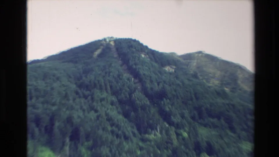 1982 New Zealand travel view of a forested mountain peak with a small summit structure silhouetted near the ridge.