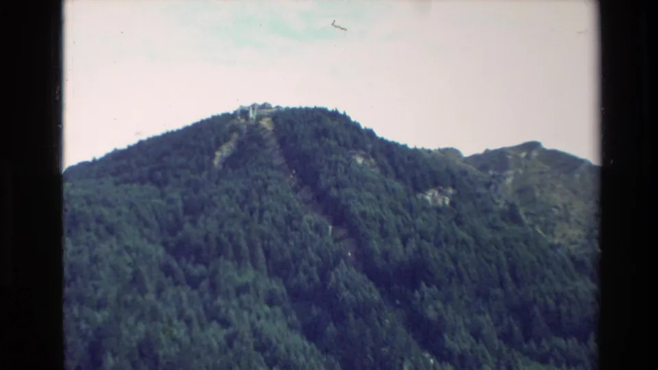 1982 New Zealand 8mm frame showing a forested mountain slope and a small distant hilltop structure beneath a pale sky.
