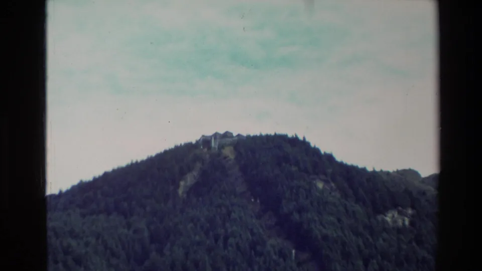 1982 New Zealand home-movie frame with a distant building set along a tree-covered mountain ridge.