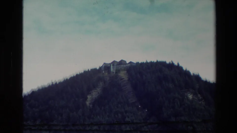 1982 New Zealand 8mm view of a hilltop structure standing above a dark evergreen-covered slope.