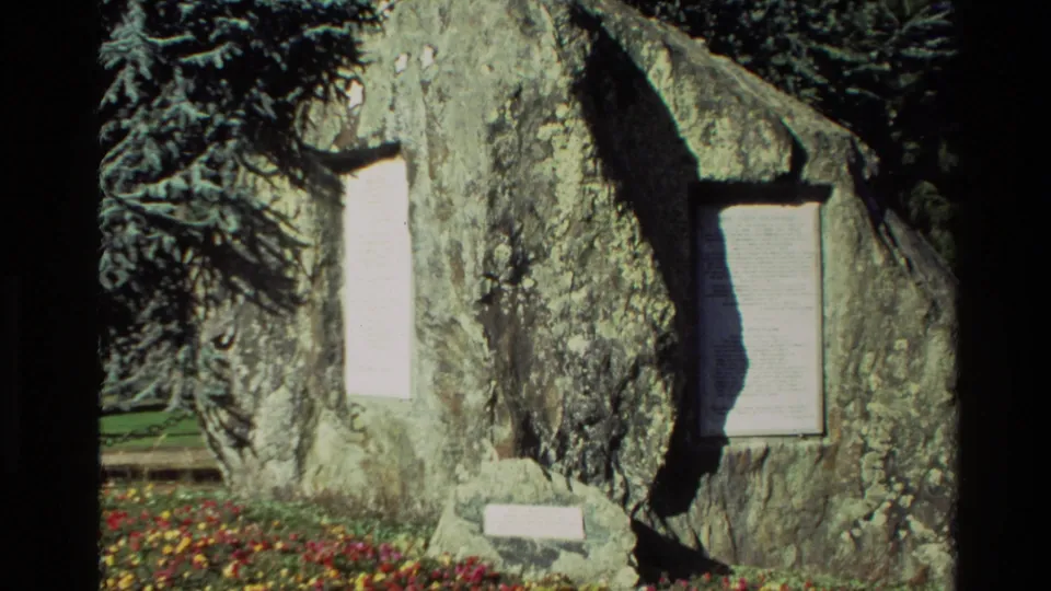 1982 New Zealand 8mm frame showing a weathered stone monument with inset plaques, framed by garden flowers and shade.