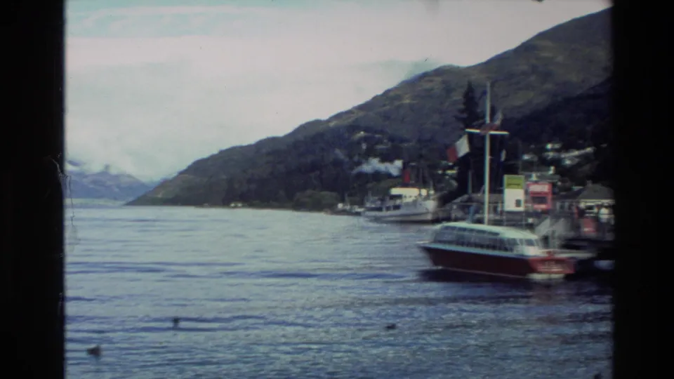 1982 New Zealand 8mm frame showing a small harbor, moored boats, calm lake water, and steep mountains under cloud.