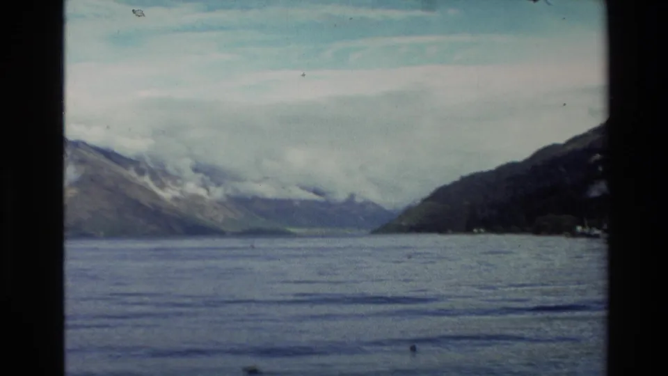 1982 New Zealand 8mm frame looking across calm lake water toward mountain slopes partly covered by low cloud.