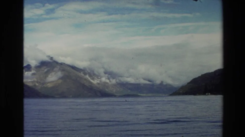 1982 New Zealand 8mm frame showing calm water, distant mountains, and low cloud stretching across the peaks.