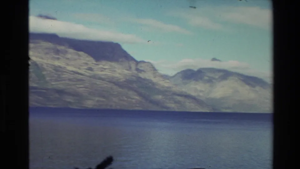 1982 New Zealand 8mm frame with a wide lake foreground and rugged mountain ridges under a soft, pale sky.