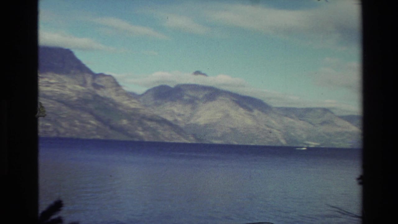 1982 New Zealand home-movie view of a calm blue lake and rugged mountains under a cloudy sky.