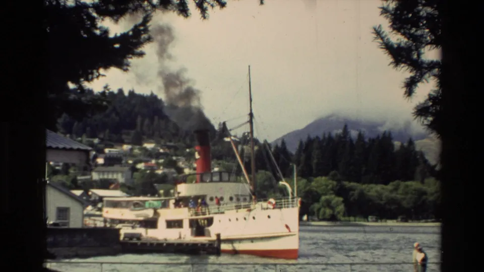 1982 New Zealand waterfront scene with a docked steamship, red smokestack, smoke plume, trees, and hillside buildings.