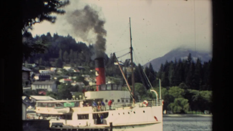 1982 New Zealand home-movie frame of a steamship at a lakeside dock, with passengers, trees, smoke, and misty hills behind.