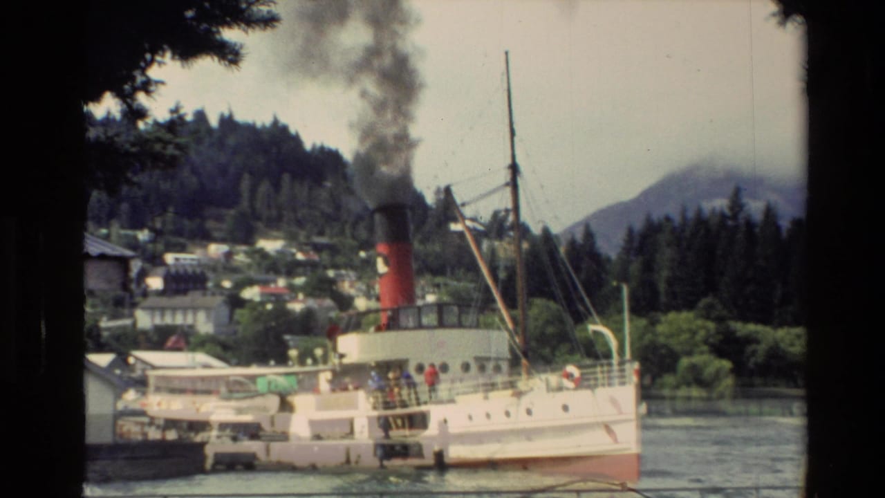 1982 New Zealand 8mm frame showing the Earnslaw steamer near shore, with black smoke, lake water, hills, buildings, and passengers on deck.